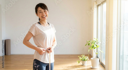 Japanese woman measuring her waist with tape measure at home, smiling confidently in bright natural light