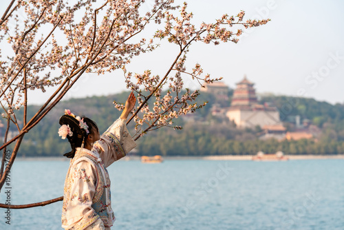 A woman in traditional Chinese dress touching a flowering cherry blossom branch on the shore of Kunming Lake with the iconic Summer Palace pagoda in Beijing
