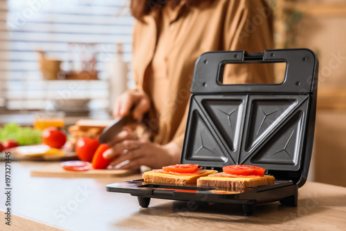 Modern sandwich maker with snacks against woman cutting tomato on table in kitchen, closeup