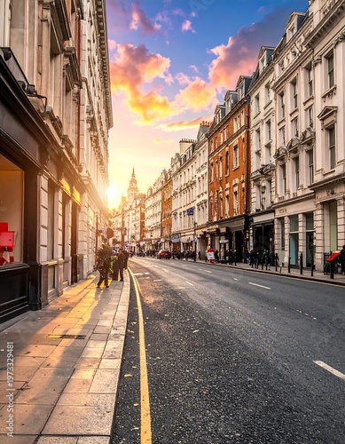 Cobblestone street scene, lined with classic architecture under a dramatic sunset sky. People walk alongside the buildings. Cars parked and driving
