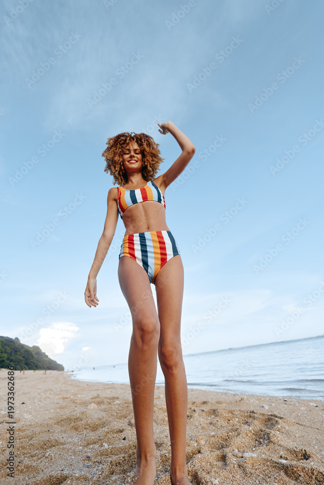 Naklejka premium Young woman in striped bikini stands on sandy beach with raised arm smiling happily woman enjoying summer vacation near calm sea water under blue sky with clouds.