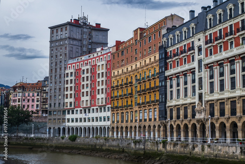Fachadas de edificios tradicionales y modernos en la Ribera del Nervión, Bilbao, mostrando sus característicos colores, balconadas y soportales junto al cauce de la ría bajo un cielo nublado.