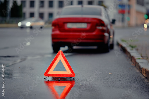 Reflective warning triangle behind broken vehicle. Car accident aftermath on dark, wet city road with emergency triangle marking hazard zone, ensuring visibility for oncoming drivers. Selective focus
