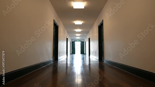Symmetrical view of a long interior hallway with white walls, multiple dark wooden doors, and a polished hardwood floor reflecting bright overhead square lights and an open doorway at the end.