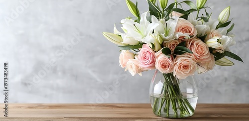 A clear glass vase holds a delicate bouquet of pale pink roses and white lilies on a wooden surface against a soft gray background.