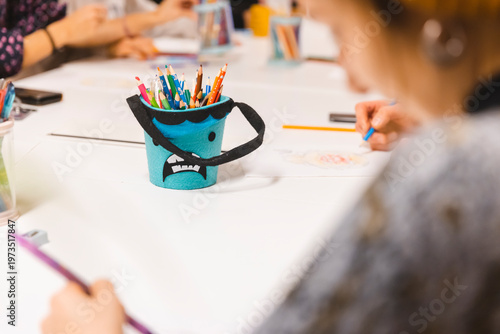 Close-up of a creative blue monster-themed pencil holder filled with colorful pencils on a white table during a children's art workshop, with blurry kids drawing and crafting in the background.