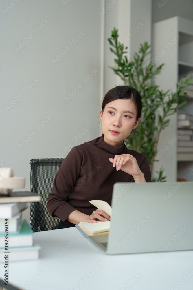 Fototapeta premium Young Asian woman student learning and thinking while studying with a laptop and books at a bright desk, focusing on online education and planning future career goals at home