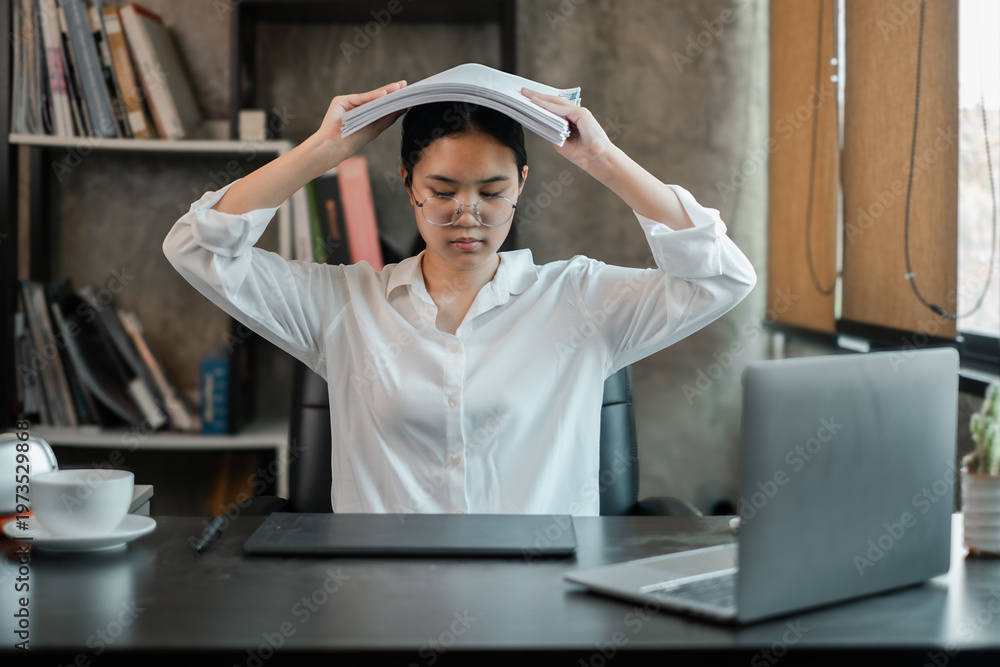 Obraz premium A young woman in an office setting appears stressed, holding documents over her head, surrounded by a laptop and books.