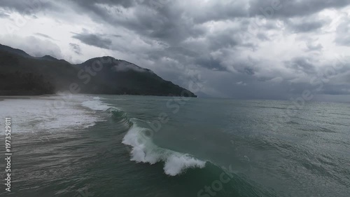 Storm waves approaching tropical coastline. Aerial drone view of rolling ocean swell under heavy clouds with mountain backdrop, creating dramatic seascape with rough water and storm atmosphere.