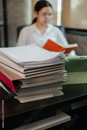 Konstfotografi A professional woman reading documents at her desk, surrounded by a stack of files in a modern office setting
