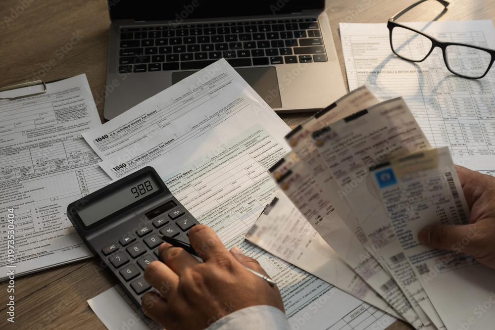 Obraz premium Close-up of a person calculating expenses with a calculator while holding multiple receipts and reviewing tax documents on a desk with a laptop, suitable for expense reporting, budgeting, reimbursemen