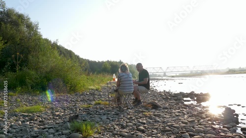 A man and a woman sit with their backs to the camera on a riverbank, relaxing and spending time outdoors