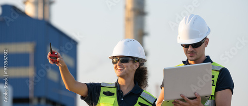 Two petroleum engineers in hardhat and safety vest reviewing blueprint and pointing at oil refinery industrial site during sunset for inspection teamwork planning discussion in energy industry.