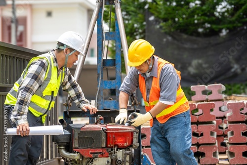Two construction workers wea safety gear inspecting equipment at outdoor construction site with bricks and greenery in background