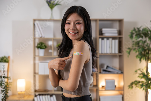 Happy asian woman showing vaccination plaster on arm smiling with proud emotion after receiving health treatment for immunity protection inside medical clinic room feeling safe