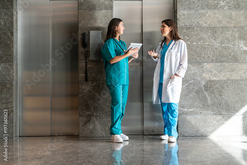 A woman in teal scrubs holds a tablet and discusses with a colleague in a white lab coat at the exit of an elevator in a hospital setting. 