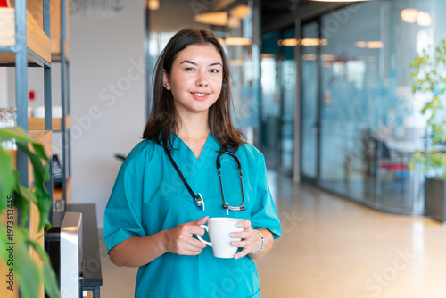 Smiling young nurse holding a cup of coffee in a hospital setting, at the camera.