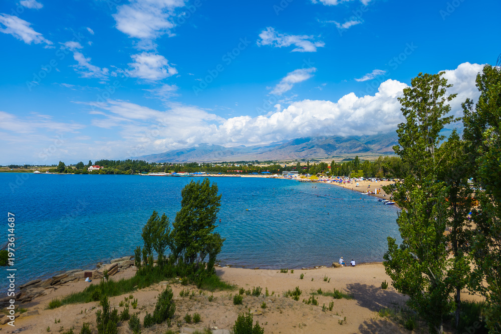 Naklejka premium Serene beach scene features clear water, distant mountains, and boats at Issyk-Kul lake. Lush trees frame the landscape, creating a peaceful summer atmosphere.
