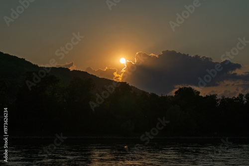 Sunset through clouds over a wooded hilly landscape.