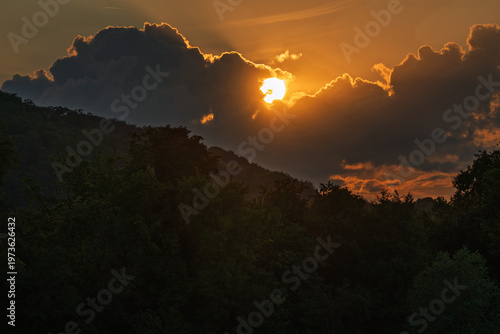 Sunset through clouds over a wooded hilly landscape.