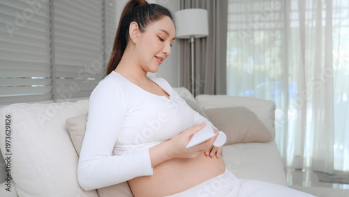 Free Download Happy pregnant woman applying stretch mark cream on her belly while sitting on a sofa. This skincare routine helps hydrate skin and improve elasticity during pregnancy for a healthy, glowing look - GFXLoad