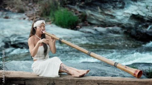 A serene moment by the river, a young woman skillfully plays a bamboo didgeridoo, showcasing her talent while surrounded by nature's beauty and soothing sounds.