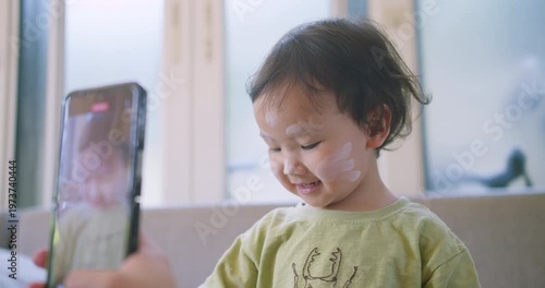 Playful Asian toddler touches white skincare cream on their face while watching a smartphone screen at home, focusing on childhood hygiene routines, digital parenting, and family memories.