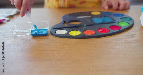 Toddler hands squeeze blue liquid from a dropper into a plastic tray beside a watercolor palette on a wooden desk, focusing on early childhood education, fine motor skills, and creative play.