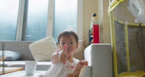 Asian toddler girl gives a thumbs up gesture over a high chair tray in a bright living room, focusing on child development, positive reinforcement, and family lifestyle.