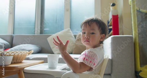 Asian toddler girl sitting looking at the camera using a white high chair in a modern living room, focusing on domestic childcare routines, early childhood development, and family mealtime.