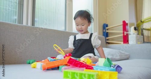 Asian toddler playfully grabs plastic building blocks using yellow toy tongs on a living room couch, focusing on early education, fine motor skill development, and independent play.