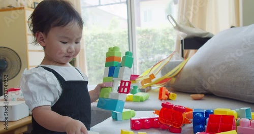 Engaged Asian toddler girl constructs a colorful tower using plastic toy blocks in a bright living room, focusing on early childhood development, creative playtime, and motor skills education.