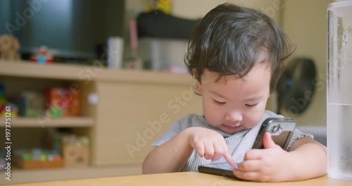 Focused Asian toddler taps and swipes a smartphone screen at a home living room table, focusing on early digital literacy, screen time engagement, and modern childhood technology habits.