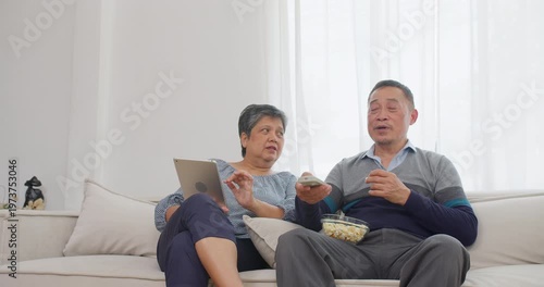 Senior Asian couple operates a television remote and digital tablet on a living room sofa, focusing on comfortable domestic leisure, active retirement lifestyle, and modern senior living.