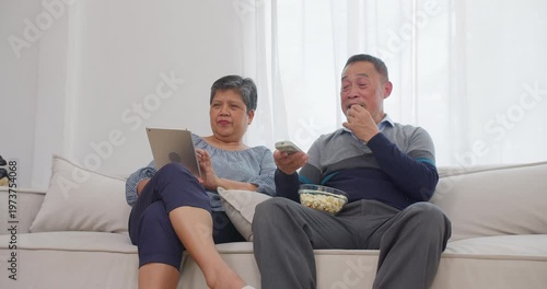 Senior Asian couple discussing entertainment options using a remote control and digital tablet on a living room sofa, focusing on active aging, domestic technology use, and joyful shared leisure.
