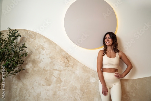Fit charismatic woman athlete leaned on gym wall in white leggings and top posing next to a empty circle add text