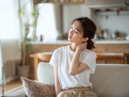 リビングのソファに座り首の痛みや肩こりに悩む表情の日本人女性