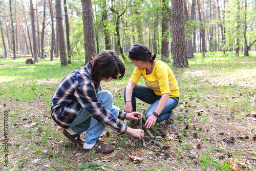 Teen son and mother building small campfire from twigs in forest clearing during outdoor family activity