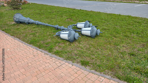 Fallen ornate gray lamppost with three lanterns lying on green grass next to a brick sidewalk