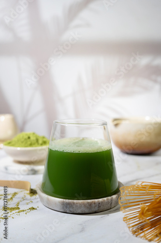Glass of green matcha tea with foam, accompanied by matcha powder, whisk and bamboo scoop.