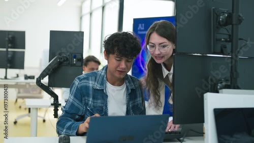 collaborative AI development team in modern tech office: young male developer coding on laptop while senior colleague leans over discussing code and data visualizations on multiple monitors	