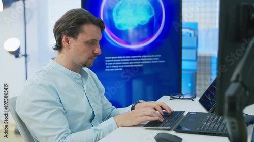 professional man code designers in modern AI development office focuses intently on coding at laptop while viewing large blue AI Assistant screen with glowing neural brain graphic, symbolizing machine
