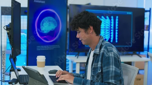 focused young male developer with curly hair in blue plaid shirt intensely codes on laptop in modern AI office, surrounded by multiple monitors displaying code neural networks and data graphs	