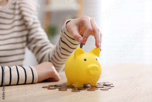 Quadro su tela Girl putting coins into piggybank at table indoors, closeup