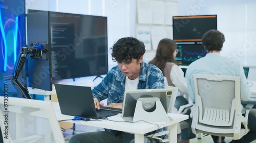focused young male developer with curly hair in blue plaid shirt intensely codes on laptop in modern AI office, surrounded by multiple monitors displaying code neural networks and data graphs	