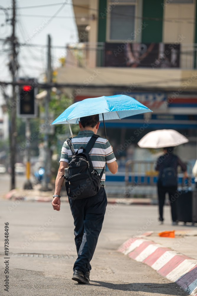 Fototapeta premium Man walking through city street holding umbrella, health safety and protection from heatstroke during heatwave, male hiding from sun under umbrella while passing by crossroad on hot summer day.