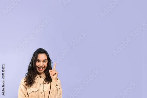 Принти на полотні A woman with long dark hair smiles and raises her index finger in a bright indoor space with a plain purple background