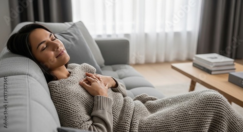 Woman resting on a cozy sofa at home. Peaceful girl relaxing with eyes closed and hands on chest. Concept of comfort, mindfulness, calm, stress relief, tranquility, lifestyle and interior apartment