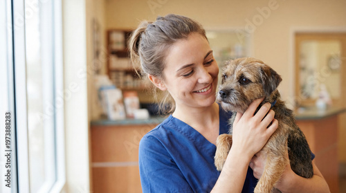 A woman in blue scrubs holds a small dog in a veterinary clinic. She smiles as she interacts with the dog, showing care and attention in her work