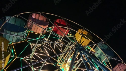 Close-up shot of a colorful Ferris wheel at night with vibrant green, yellow, and orange lights against a dark background